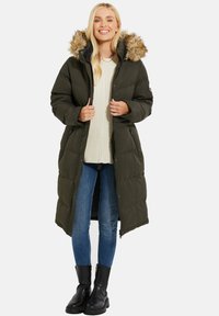 Parka in un verde scuro imbottito con bordatura in pelliccia sintetica, chiusura frontale con zip e tasche laterali, indossata sopra un maglione chiaro e jeans blu. Stivali neri alla caviglia.