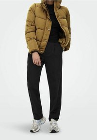 Brązowa kurtka puchowa z zamkiem błyskawicznym z przodu i zapięciem na zatrzaski, w zestawie z czarnymi spodniami i wielokolorowymi szarymi trampkami.