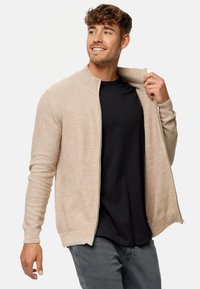 Cárdigan beige con cremallera y diseño de punto texturizado, cuello alto y puños acanalados, llevado sobre una camiseta negra y jeans grises.