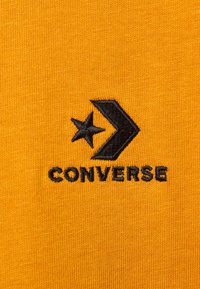 Gult bomullstyg med en texturerad yta har en svart broderad "Converse"-logotyp och en stjärna med en pil-design.