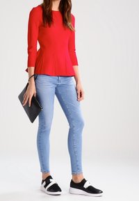 Haut peplum rouge avec un motif texturé, associé à un jean skinny bleu clair et des baskets noires à enfiler avec des straps blancs.