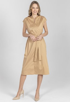 Uśmiechnięta kobieta o falowanych blond włosach ma na sobie beżową sukienkę midi z krótkimi rękawami, paskiem w talii oraz beżowe szpilki, stoi na tle szarości.