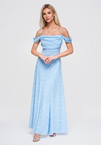 Femme blonde portant une robe de soirée bleu clair épaules dénudées avec fines bretelles et des sandales argentées à talons hauts, debout, les mains jointes.
