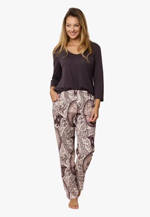 Kobieta stojąca boso, ubrana w ciemną bluzkę z długim rękawem i jasne spodnie w wzór paisley, z rękami w kieszeniach, uśmiechająca się.