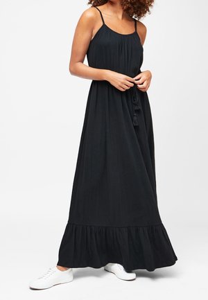 Femme portant une robe maxi noire sans manches avec de fines bretelles et des baskets blanches, debout devant un fond uni.