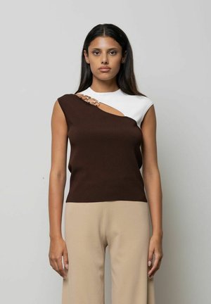 Donna con capelli lunghi e scuri che indossa un top asimmetrico senza maniche con sezioni bianche e marroni e pantaloni beige, in piedi davanti a uno sfondo semplice.