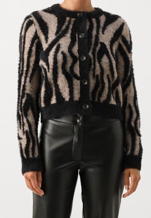Mujer con un cárdigan de rayas de cebra borroso en negro y beige con botones, combinado con pantalones de cuero negros de talle alto.