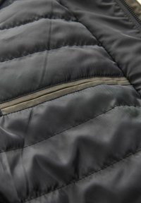 Jachetă neagră matlasată cu un model în dungi orizontale, textură netedă și o buzunar cu fermoar gri contrastant de-a lungul cusăturii.