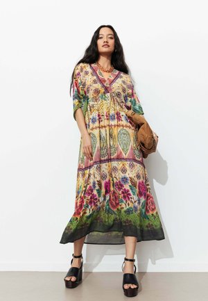 Femme portant une robe midi à motifs floraux et géométriques colorés, des sandales à plateforme noires, un collier en or, tenant une veste en daim marron.