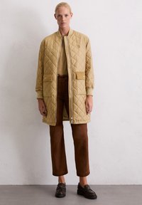 Une femme se tient debout, portant un long manteau matelassé beige, un pantalon marron, un pull beige et des mocassins marron foncé, contre un mur clair texturé.