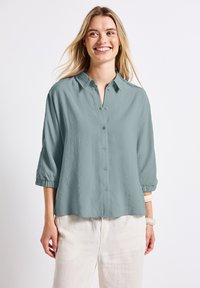 Camicia button-up azzurro chiaro con colletto, maniche a tre quarti e vestibilità rilassata. Tessuto morbido e texture. Indossata con pantaloni di colore chiaro.