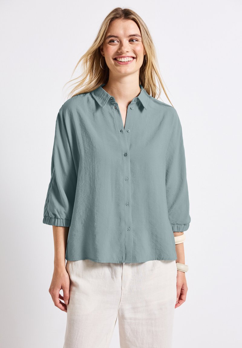 Camicia button-up azzurro chiaro con colletto, maniche a tre quarti e vestibilità rilassata. Tessuto morbido e texture. Indossata con pantaloni di colore chiaro.