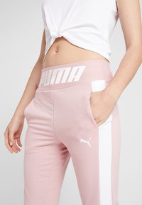 Ljusrosa joggerbyxor med ett vitt band och logotyp. Har sidofickor och ett brett elastiskt midjeband med fet "PUMA"-text. Mjuk textur.