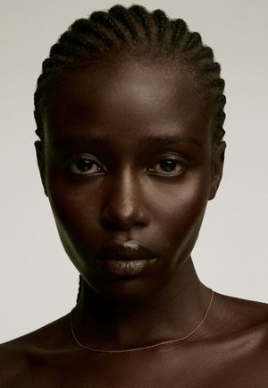 Primer plano de una persona con cabello trenzado y piel oscura, con una textura suave y un brillo sutil, que lleva un collar metálico delgado.