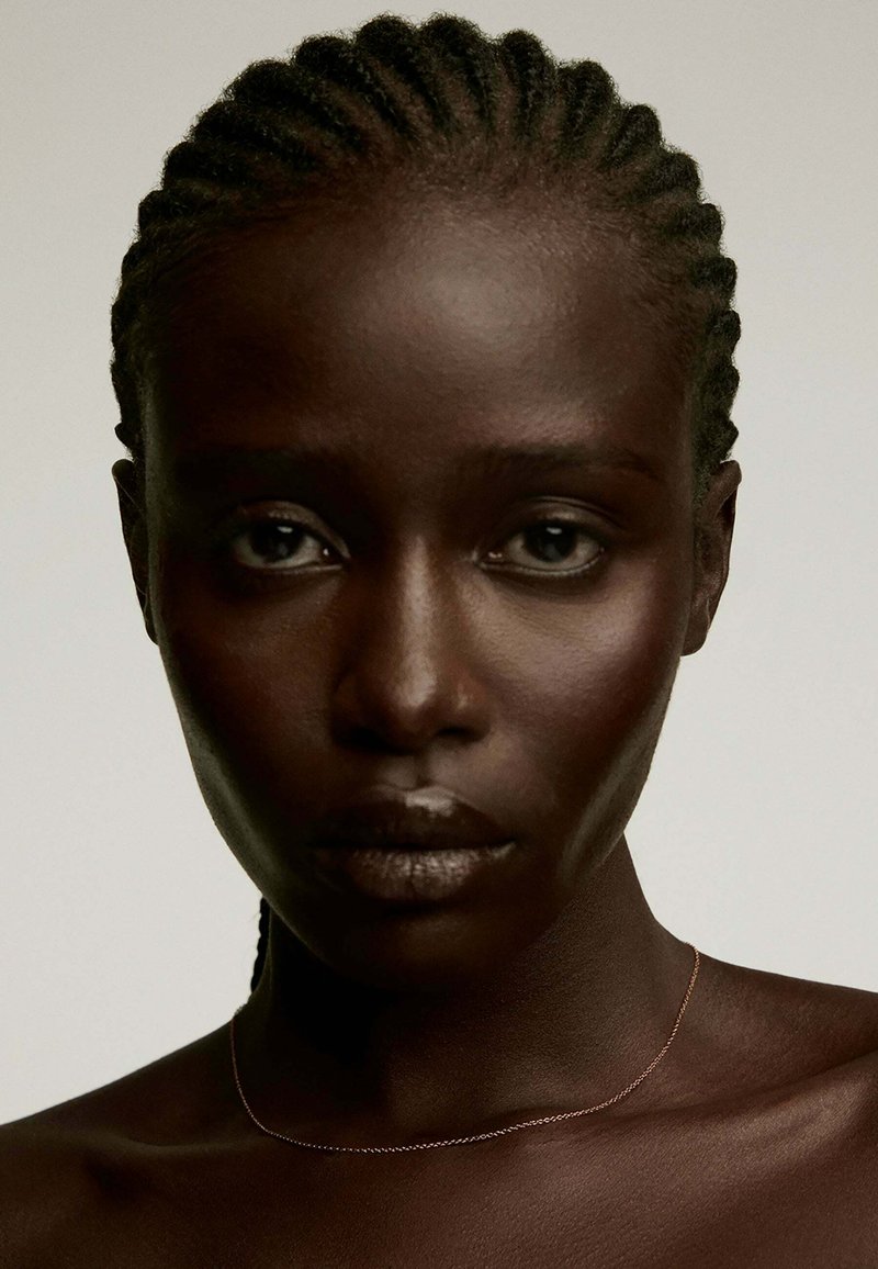 Primer plano de una persona con cabello trenzado y piel oscura, con una textura suave y un brillo sutil, que lleva un collar metálico delgado.
