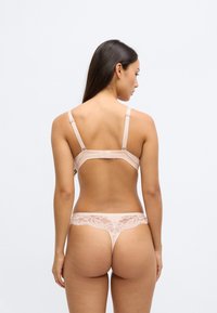 Zestaw bielizny w kolorze nude wykonany z gładkiego, elastycznego materiału, ozdobiony koronkowymi detalami i regulowanymi ramiączkami. Figi typu string z niskim stanem z tyłu.