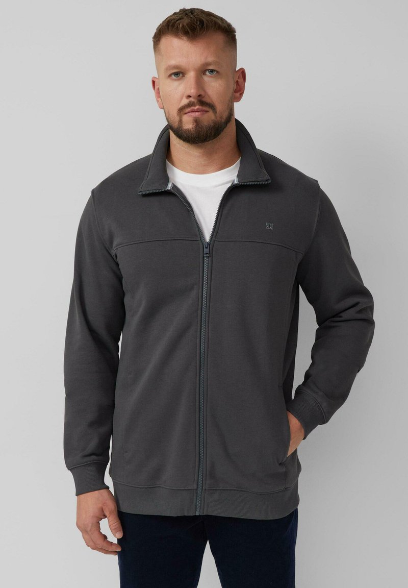 Giacca a zip grigio scuro in morbido tessuto, con colletto alto, polsini a costine e tasche laterali. Design semplice con minimi dettagli.