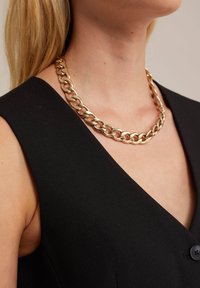 Auksiniu tonu grandinėlė, turinti tarpusavyje susijungusias jungtis, dėvima virš juodos be rankovių palaidinės su V formos iškirpte. Lygus, poliruotas paviršius.