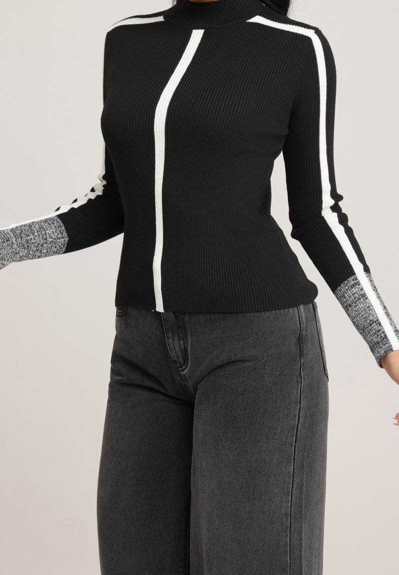 Femme portant un pull noir côtelé avec des rayures blanches verticales et des manches grises en bloc de couleur, associé à un jean taille haute gris foncé.