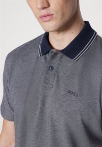 Tumši pelēks polo krekls ar tumši zilu apkakli, kas rotāta ar baltām svītrām, ar vienu pogu un nelielu logo uz krūtīm. Struktūrēta auduma.