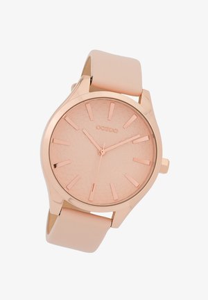 Montre en or rose avec un cadran texturé, des index horaires minimalistes et un bracelet en silicone lisse de couleur rose pâle. Design épuré et moderne.
