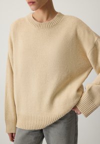 Bežový pletený sveter s rebrovaným okrúhlym výstrihom, vo voľnom strihu a rebrovanými manžetami. Textúrovaná tkanina s jemným vzhľadom. Skombinovaný so šedými džínsami.