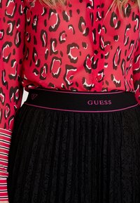 Leopardprint gennemskinnelig pink bluse med en sort blonde nederdel. Den sorte elastiske talje har "GUESS" logoet i pink. Stribet ærmedetalje.