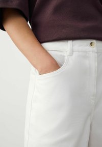 Pantaloni di cotone bianchi con una superficie liscia, dotati di una tasca laterale e di un bottone visibile in vita, abbinati a un top di un viola scuro.