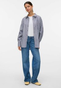 Camicia a righe blu e bianca, abbinata a jeans a gamba larga blu e scarpe gialle. La camicia ha una vestibilità rilassata e tasca sul petto.