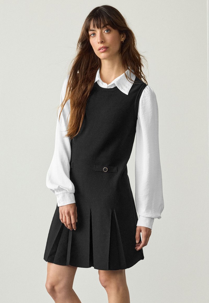 Femme aux longs cheveux bruns portant une robe noire plissée sans manches par-dessus une blouse blanche à manches longues avec col, debout devant un fond uni.