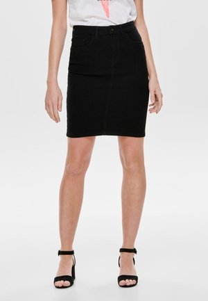 Femme portant une jupe noire mi-longue, un t-shirt blanc avec un motif graphique et des sandales noires à talons hauts avec brides à la cheville, debout devant un fond uni.
