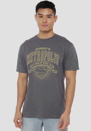 Young man wearing a gray T-shirt with "University of Metropolis Man of Steel" and a Superman logo in yellow print, paired with light blue jeans.