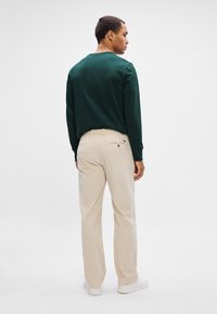 Mężczyzna stojący tyłem, ubrany w ciemnozielony sweatshirt, beżowe spodnie sztruksowe i białe trampki na jednolitym białym tle.