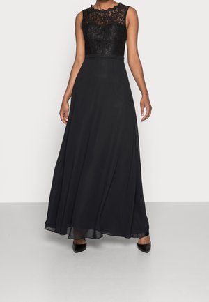 Femme portant une robe de soirée noire sans manches avec un corsage en dentelle et une jupe fluide en chiffon, assortie à des escarpins noirs à bout pointu.