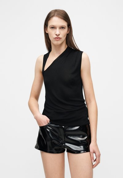 Γυναίκα με μαύρο ασύμμετρο τοπ χωρίς μανίκια και γυαλιστερά μαύρα δερμάτινα σορτς, στέκεται με το ένα χέρι στην τσέπη της μπροστά σε λευκό φόντο.