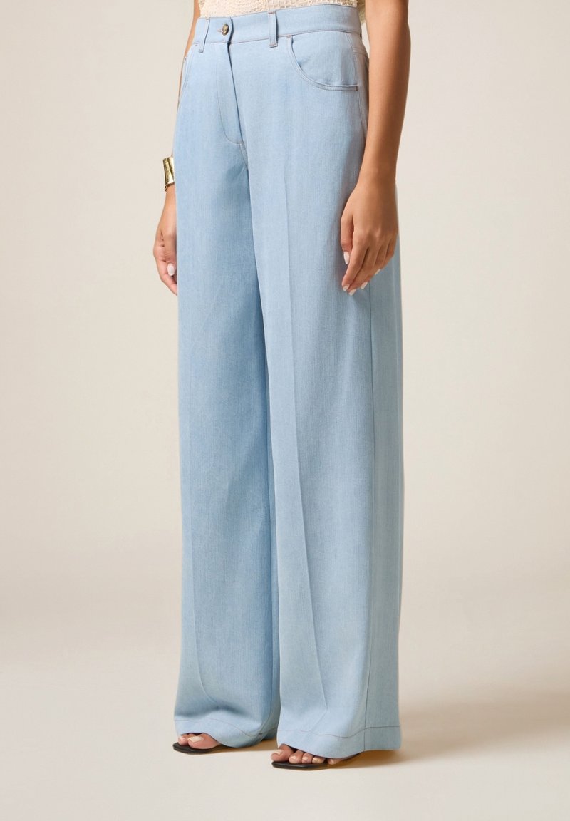 Pantalon large bleu clair porté par une personne debout avec un haut beige et des sandales noires à bout ouvert, sur un fond uni.
