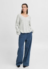 Grau gestreifter Strickpullover mit weitem Halsausschnitt und langen Ärmeln, kombiniert mit blauen, gestreiften weit geschnittenen Hosen und schwarzen spitzen Schuhen.