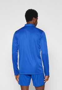 Μακρυμάνικο αθλητικό πουκάμισο σε φωτεινό μπλε χρώμα, κατασκευασμένο από λείο, ελαφρύ ύφασμα. Διαθέτει γιακά και εφαρμοστή γραμμή, συνδυασμένο με σορτς στο ίδιο μοτίβο.