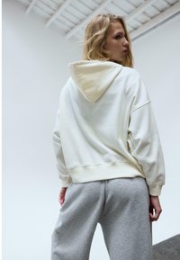 Sweat à capuche surdimensionné couleur crème avec une capuche, présentant un tissu texturé et des poignets côtelés, associé à un pantalon de survêtement gris.