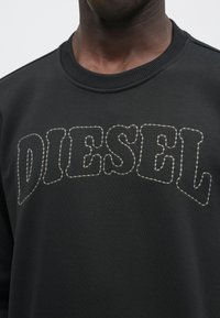 Μαύρο φούτερ με ριγωτό στρογγυλό λαιμό, διακοσμημένο με το κέντημα "DIESEL" σε ανοιχτό γκρι νήμα. Το υλικό είναι απαλό και υφασμένο, με άνετη, χαλαρή εφαρμογή.