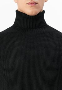 Man met een zwarte geribbelde coltrui, die een gestructureerd breipatroon laat zien, met een close-up van de nek en de kin.