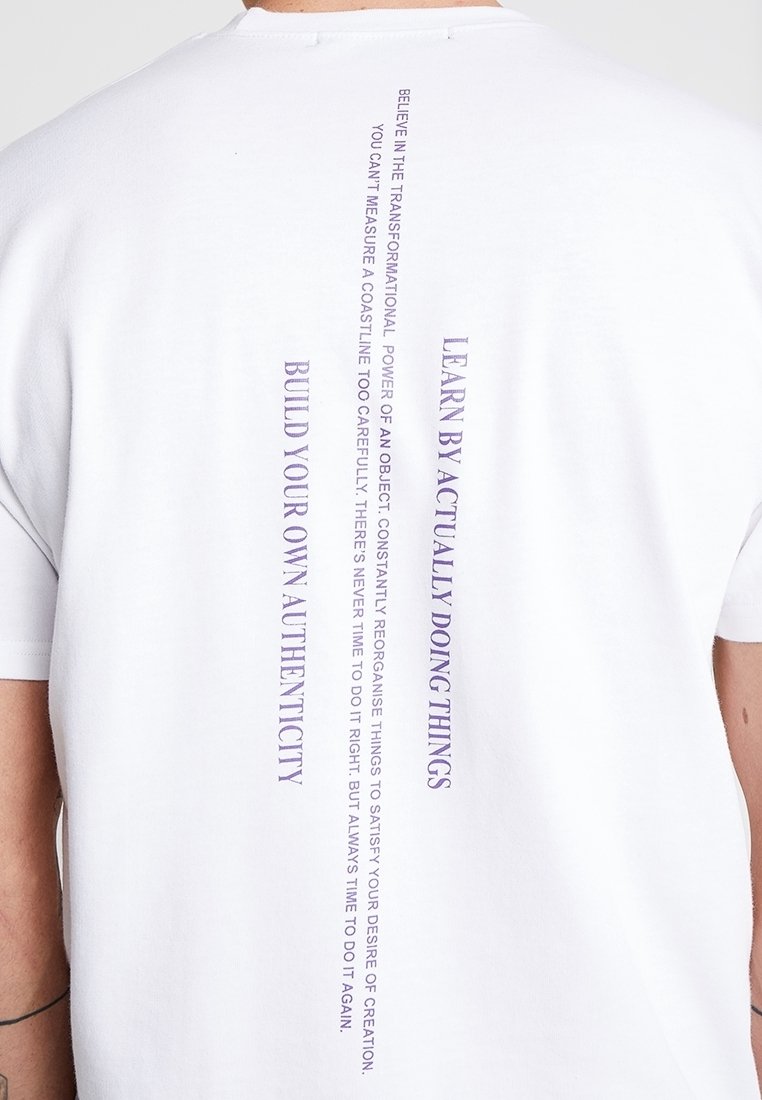 Bílá tričko s vertikálním fialovým textem: "UČ SE SKUTEČNÝM DĚLÁNÍM VĚCÍ" a "VYBUĎ SI SVOU VLASTNÍ AUTENTICITU" na zádech.