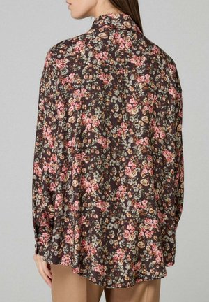 Donna che indossa una camicia marrone scuro a maniche lunghe, larga, con un piccolo motivo floreale rosa, bianco e arancione, abbinata a pantaloni beige.