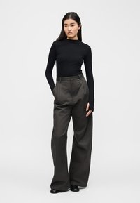 Γυναίκα με μακριά μαύρα μαλλιά, να φοράει εφαρμοστή μαύρη μπλούζα με μακριά μανίκια και φαρδιά, ψηλόμεση σκουρόχρωμη γκρι παντελόνα, στέκεται με τα χέρια στις τσέπες.