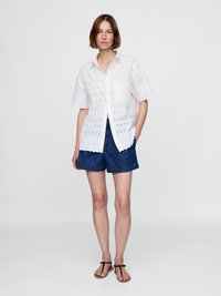 Hvit bomullsskjorte med øyelet detaljer, korte ermer og knappelukking, sammen med blå denimshorts og svarte sandaler.