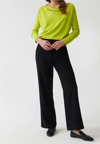 Maglione verde brillante con scollatura a barchetta, abbinato a pantaloni neri a gamba larga e mocassini neri. Trame morbide, design casual.