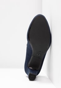 Chaussure pour femme en daim bleu marine positionnée sur son talon, montrant une semelle noire lisse avec la taille 37 et les marquages du logo de la marque.