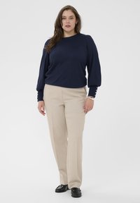 Navyblauwe trui met pofmouwen, gecombineerd met beige op maat gemaakte broeken. Zwarte instapschoenen en een zilveren armband maken de look compleet.