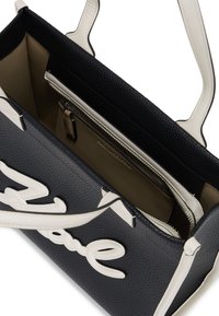 Geantă tote din piele neagră cu un finisaj texturat, dotată cu mânere albe și detalii cu script de semnătură în față. Interiorul include un buzunar cu fermoar.