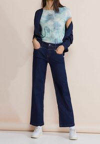 Kvinna i en ljusblå mönstrad t-shirt, marinblå jacka som hänger över axlarna, mörkblå vida jeans och vita sneakers.